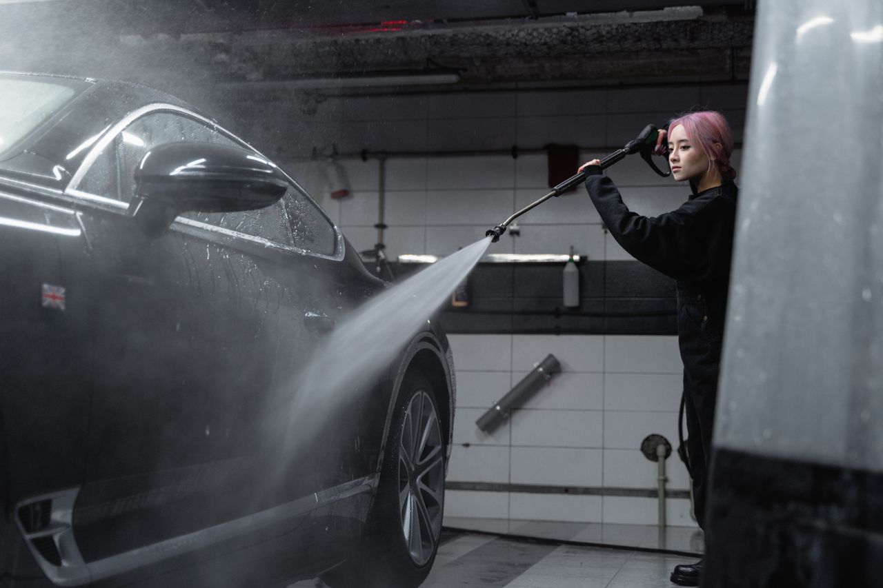 who-we-are Woman power washing a black car in an indoor garage using a high-pressure washer.
