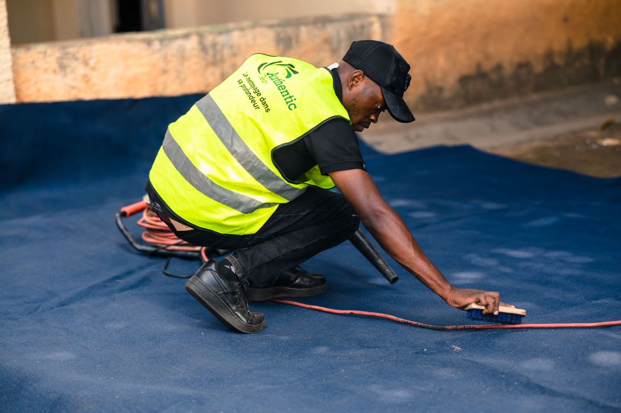 services-03 A worker dressed in a safety vest cleans a blue carpet outdoors with a brush.